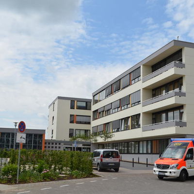 Das Foto zeigt die Frontansicht des viergeschossigen St. Rochus-Krankenhauses mit der kleinen Kapelle. Im Vordergrund steht ein Rettugswagen.
